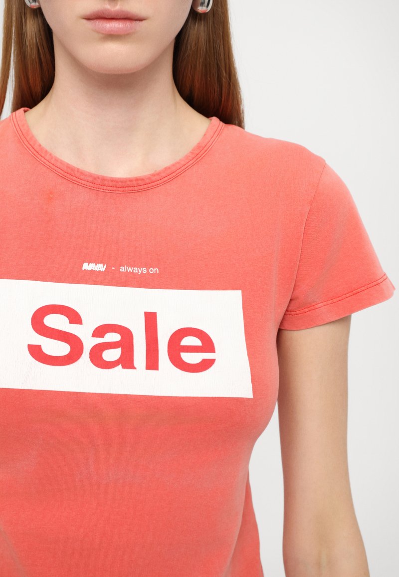 Camiseta coral con mangas cortas y cuello redondo, que presenta un parche rectangular blanco con el texto rojo "Oferta" y texto adicional arriba.