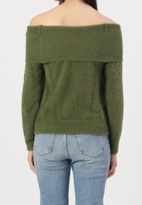 Maglione verde off-the-shoulder con texture morbida e pelosa, colletto a costine, maniche lunghe e fondo aderente, abbinato a jeans azzurri chiari.