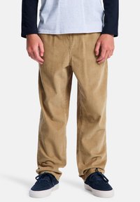 Beige corduroy broek met verticale ribbels, rechte pijpdesign, lichte textuur, gecombineerd met donkerblauwe sneakers en een grijs overhemd met lange mouwen.