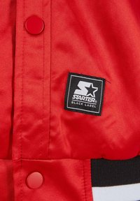 Rød satinjakke med en sort "Starter Black Label"-patch og en rund rød trykknap. Struktureret stof med sorte og hvide stribede detaljer.