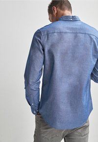 Man in een lichtblauwe overhemden met lange mouwen en een grijze broek, gezien van achteren tegen een lichte achtergrond.