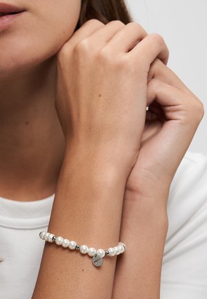 Woman wearing white beaded bracelet with silver accents and small round charm on wrist against a plain background.