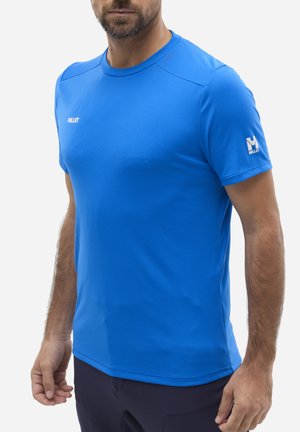 Mann trägt ein blaues Millet Sport-T-Shirt mit kurzen Ärmeln und Logos auf Brust und Ärmel, steht vor einem weißen Hintergrund.