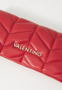 Rdeča prešita usnjena torbica s svetlečim zlatim logotipom "Valentino". Opremljena je z vzorcem chevron in gladko teksturo. Kompaktna pravokotna oblika.