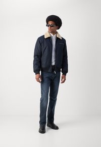 Chaqueta bomber navy con un collar de piel sintética beige, combinada con una camisa clara y jeans azul oscuro. Botas negras completan el atuendo.