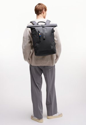 Henkilö, jolla on päällään beige neule ja harmaat housut, kantaa selässään suurta mustaa rullattavaa reppua, jossa lukee "GOT BAG".