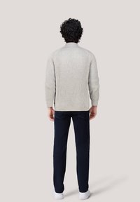 Maglione a coste grigio chiaro con colletto alto, indossato sopra pantaloni blu scuro e sneakers bianche, mostrato di spalle.