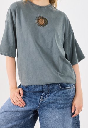 Osoba nosząca wyblakły szary, oversize T-shirt z emblematem słońca i księżyca, w połączeniu z luźnymi, wysokim stanem niebieskimi dżinsami, ręka spoczywa na biodrze.