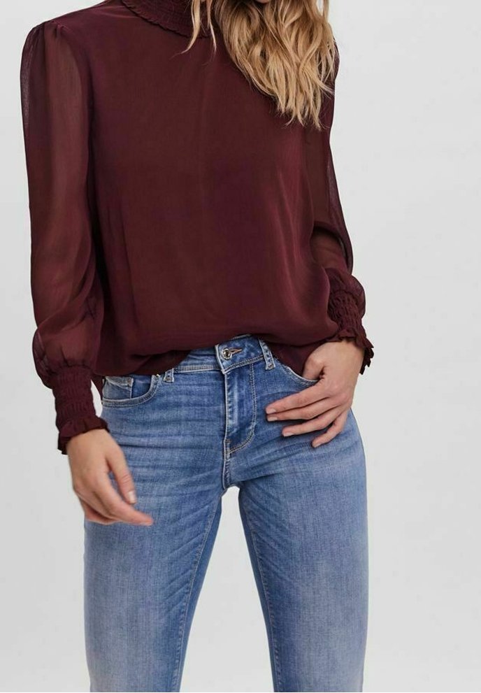 Femme portant un chemisier bordeaux transparent à manches longues avec des poignets et un col à volants, associé à un jean bleu clair taille haute.