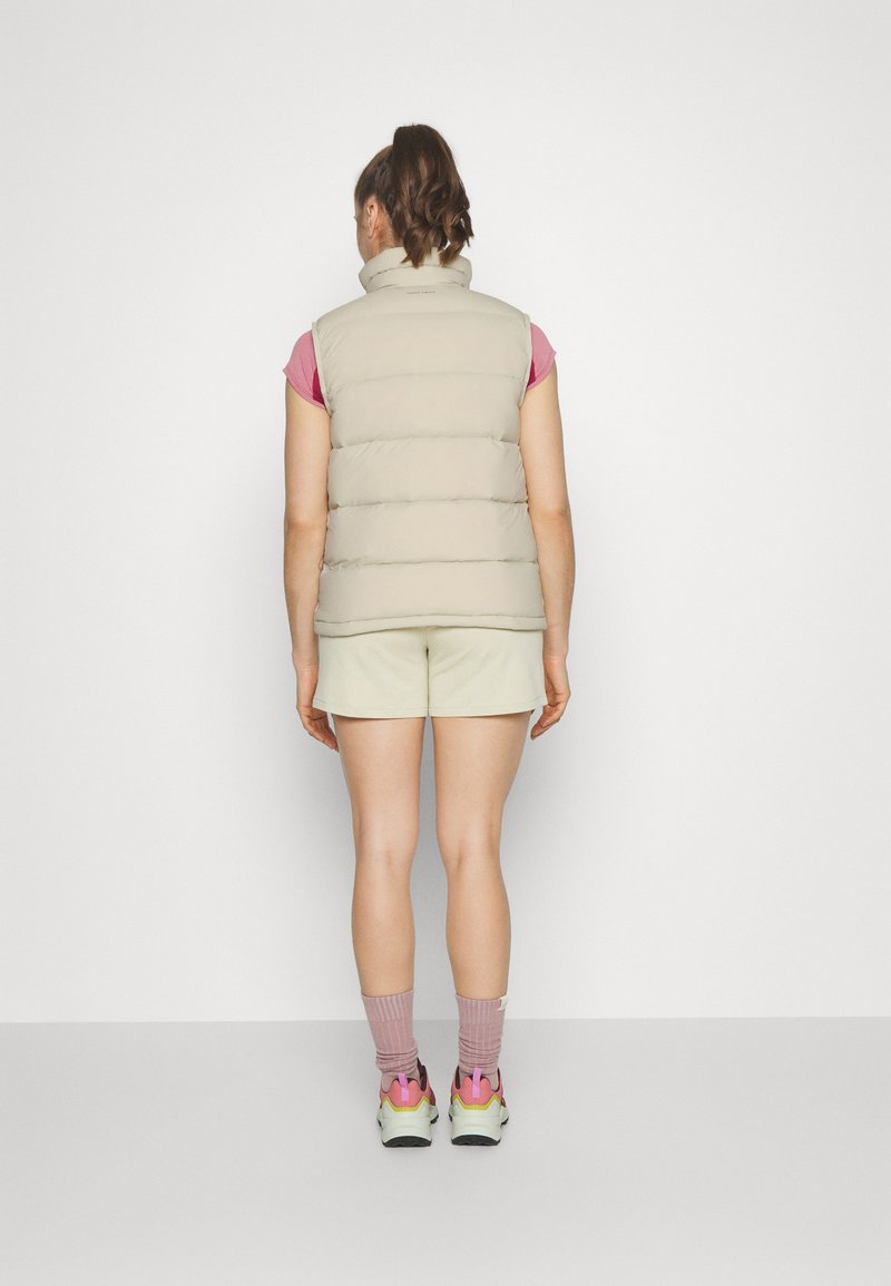 Beige Steppweste mit hohem Kragen und kurzen Ärmeln. Getragen über einem pinken Shirt, kombiniert mit hellen Shorts und bunten Sneakers.