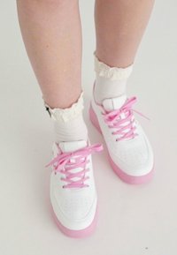 Baskets blanches avec une semelle rose, ornées de lacets roses et de détails perforés. Portées avec des chaussettes blanches côtelées et des poignets à volants.