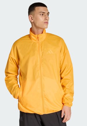 Keltainen kevyt takki, jossa on vetoketjullinen etuosa, korkea kaulus ja joustavat hihansuut. Vasemmassa yläkulmassa hienovarainen logo.