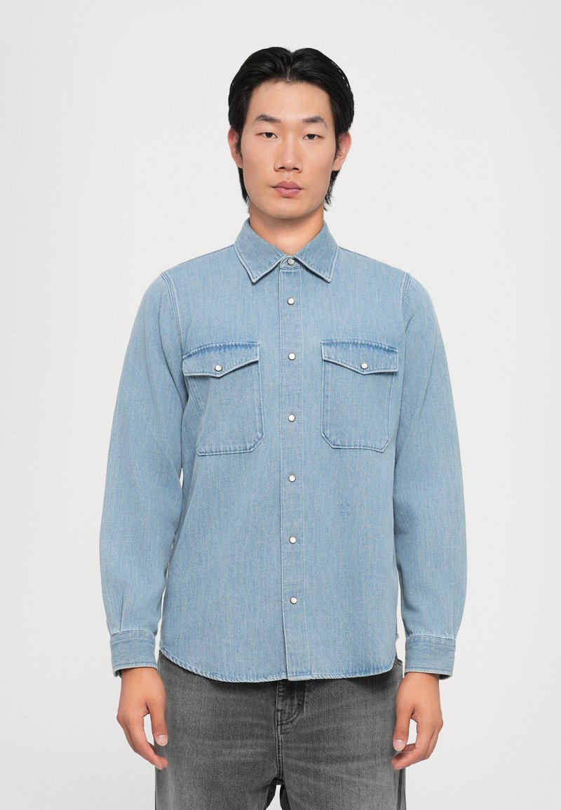 Lyseblå denimskjorte med lange ermer, knappelukking, to brystlommer, kontrastsømbånd og spiss krage.