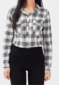 Chemise à carreaux noir et blanc en tissu doux avec manches longues, deux poches avant et un col pointu. Associée à un pantalon noir.