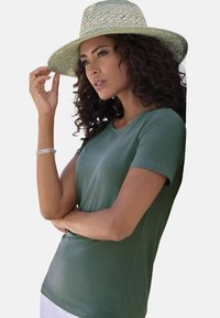 T-shirt verde a maniche corte realizzato in un tessuto morbido. La modella indossa anche un cappello di paglia intrecciato con una larga tesa e tocca la tesa.