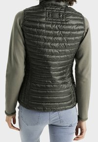 Mörkgrön isolerad väst med quiltad textur, hög krage och en åtsittande passform, lager över en ljusgrå långärmad tröja.