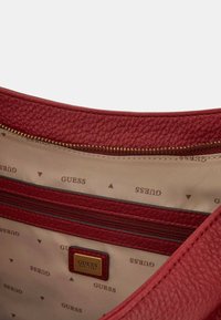 Interno di una borsa in pelle rossa con una cerniera dorata, foderato in beige con stampa del logo 'GUESS' e un piccolo etichetta in pelle con i dettagli del marchio.