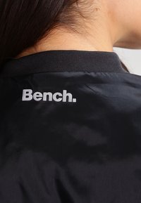 Czarna kurtka o gładkiej, błyszczącej fakturze z czarnym prążkowanym kołnierzem. Na plecach widnieje biała logo "Bench." w pogrubionej czcionce.