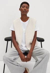 Beige Fleecepulli mit durchgehendem Reißverschluss, getragen über einem weißen T-Shirt. Das Model sitzt auf einem Stuhl und trägt graue Hosen mit weitem Bein und beige Schuhe.