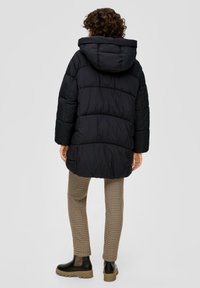 Zwarte pufferjack met capuchon, met een gestikt ontwerp en zichtbare stiksels. Gecombineerd met geruite beige broek en zwarte enkellaarsjes.