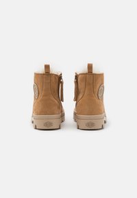 Botine din suede maro, cu o căptușeală pufoasă albă, detalii cu fermoar și talpă din cauciuc. Prezintă un logo circular pe partea exterioară.