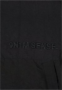 Fekete szövet, textúrált felülettel, amelyen fekete cérnával hímzett "9N1M SENSE" felirat található. Az egész design minimalista, világos minták nélkül.
