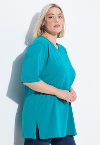 T-shirt oversize teal con maniche corte, scollo rotondo e spacchi laterali, realizzato in tessuto morbido, indossato con jeans blu scuro.