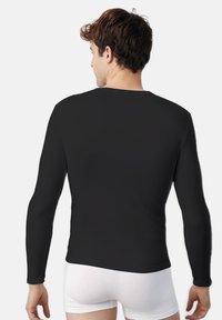 Camisa de manga larga negra con cuello redondo. Textura suave, diseño ajustado y sin patrones. Llevada con ropa interior blanca, vista desde la parte de atrás.