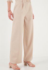 Beige wide-leg pants with a smooth texture, high waist, two large buttons, and side pockets. Worn with strappy heels.