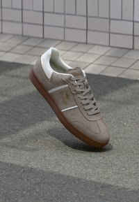 Graue Wildledersneaker mit weißen Lederakzenten, strukturiertem weißen Zungenbereich, Schnürsenkeln und einer braunen Gummisohle; verfügt über ein Logo an der Seite.
