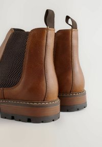 Barna bőr Chelsea csizmák fekete elasztikus oldalsó panelek, húzófülek és vastag profilú talpakkal világos háttér előtt.