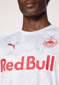 Hvid sportsjersey med et struktureret design, rød "Red Bull" skrift, en rød Puma-logo og et FC Salzburg-mærke på brystet.