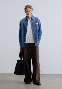 Πουκάμισο από τζιν με κουμπιά, λευκή εσωτερική μπλούζα, καφέ παντελόνι με φαρδιά μπατζάκια, μαύρη τσάντα tote και μαύρα παπούτσια. Ουδέτερο φόντο με γκρι πάτωμα.