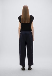 Zwart gebreide top met kapmouwtjes, gecombineerd met losse marineblauwe broek en zwarte schoenen. Het haar is stijl en lang, persoon kijkt van de kijker af.