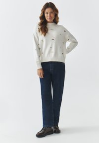 Crème gebreide trui met meerkleurige borduursels, hoge hals en ribgebreide manchetten, gecombineerd met donkere denim jeans en bruine veterschoenen.