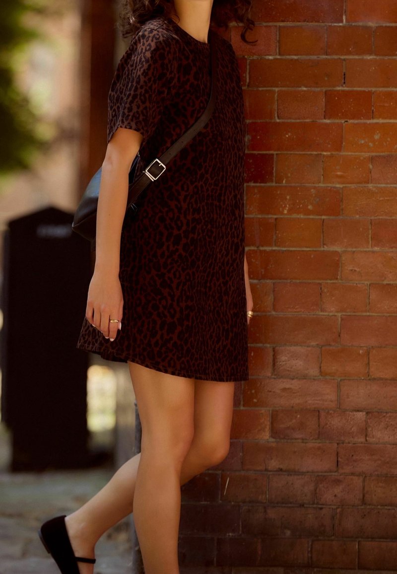 Femme portant une courte robe à motif léopard et des chaussures noires, debout près d'un mur en briques rouges avec un sac bandoulière noir.