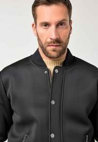 Giacca bomber nera con collo a coste. Presenta bottoni a pressione argentati, tasche con cerniera e un tessuto liscio e strutturato.