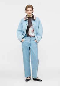 Άτομο που φοράει ανοιχτό μπλε τζιν μπουφάν με πολύχρωμο γιακά, μαύρο πουκάμισο με λουλουδάτο μοτίβο, τζιν παντελόνι που ταιριάζει και μαύρα επίπεδα παπούτσια με λουράκια γύρω από τον αστράγαλο.