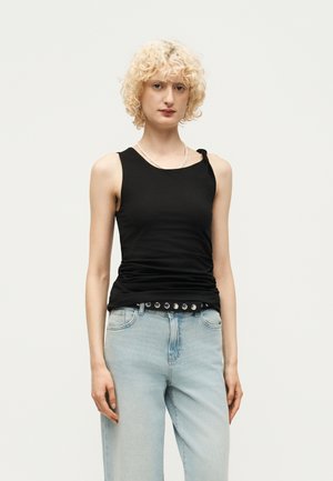 Jeune personne aux cheveux blonds bouclés portant un haut noir sans manches avec un détail nœud et un jean bleu clair sur un fond uni.