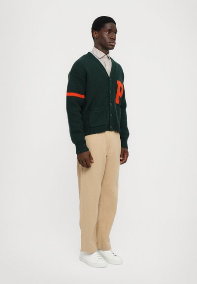 Cárdigan de punto verde oscuro con acentos naranjas, que presenta una gran "P" en el pecho, cierre de botones y bolsillos delanteros, combinado con pantalones beiges y zapatillas blancas.