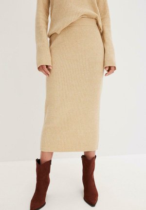 Maglione a coste beige con maniche lunghe e una silhouette aderente, abbinato a una gonna midi coordinata, anch'essa con maniche lunghe e silhouette aderente, il tutto completato da stivali chelsea in suede marrone.