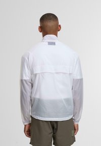 Mand med kort hår iført en hvid langærmet jakke og olivengrønne shorts, stående med ryggen til mod en ensfarvet baggrund.