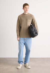 Young man wearing beige ribbed sweater, light blue jeans, beige sneakers, holding black leather duffel bag against plain white background.