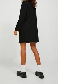 Svart långärmad klänning med rak skärning, knä-lång, med en slät textur. Parad med svarta skor och vita strumpor.