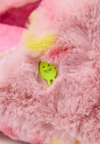 Zapatilla esponjosa de color rosa con acentos amarillos y un pequeño icono de cara sonriente verde asomándose a través del material. Textura suave con colores vibrantes.