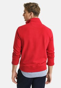 Rode sweatshirt met een hoge kraag, geribbelde manchetten en zoom. Gelaagd over een blauw-wit gestreept shirt met kleine patronen zichtbaar.