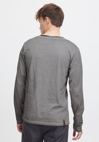 Langarmshirt aus dunkelgrauem Stoff mit lockerem Schnitt, Rundhalsausschnitt und einem braunen Logotag auf der unteren linken Seite. Glatte Textur.