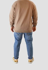 Hombre de pie de espaldas, vistiendo un sudadera marrón, jeans azules y zapatillas beige, contra un fondo claro y liso.