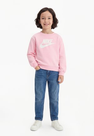 Child with shoulder-length dark hair wearing a pink Nike sweatshirt, blue jeans, and white sneakers, standing with one hand in pocket.