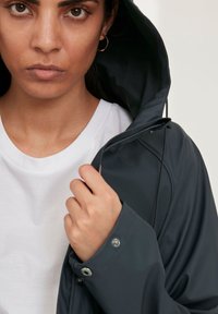 Donkergrijze hoodie met een gladde textuur, volledig rits, en drukknoopmanchetten. G gedragen over een witte T-shirt. Close-up van het gezicht.
